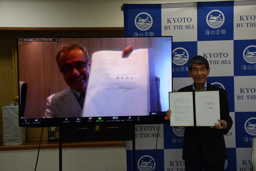 オンライン協定調印式で協定書を掲げるJapan-sanのHuw Williams代表（左）と海の京都DMOの森屋松吉社長